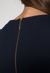 Mörkblå klänning i ett mjukt tyg, med en guldfärgad dragkedja på baksidan som detaljerar stängningen och ger kontrast mot materialet.