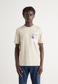 Beige bomull t-skjorte med et brystmotiv i svart og hvitt, rund hals og korte ermer, stylet med blå denimjeans.