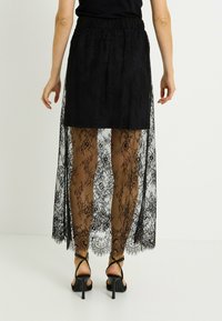 Jupe longue en dentelle noire avec un design en couches. Superposition en dentelle noire transparente et jupon noir uni. Dotée d'une taille froncée.