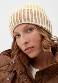 De gebreide muts heeft afwisselende gele, beige en crème strepen. Het model draagt een bruine pufferjack met drukknoop- en ritsdetails.