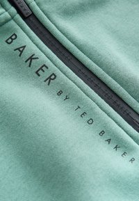 Nahaufnahme eines grünen Stoffes mit schwarzem Text ""BAKER BY TED BAKER"" nahe einem schwarzen Reißverschluss.