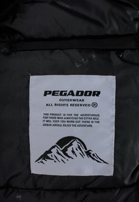 Melnā apģērba etiķete ar treknu tekstu "PEGADOR" un kalnu ilustrāciju. Audums ir gluds, un etiķete ir taisnstūrveida.