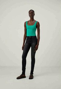 Πράσινο εφαρμοστό τι-σέρτ, σκούρο skinny τζιν και μαύρα πέδιλα. Απλός σχεδιασμός; λεία υφή; χωρίς ορατά μοτίβα ή διακοσμητικά στοιχεία.