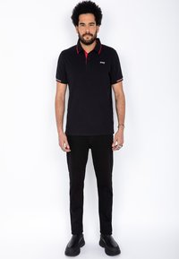 Svart polo-skjorte med røde detaljer på kragen og ermeendingene, kombinert med svarte bukser og svarte støvler. Enkel, tettsittende design.