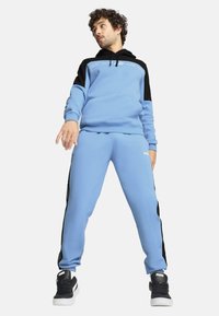 Mėlynai padengta hoodie su juodais akcentais, derinama su atitinkamais mėlynais džinsais su šoninėmis juostelėmis. Juodi sportbačiai užbaigia aprangą.