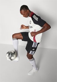 Voetbalkleding met een wit shirt met zwarte en kastanjebruine accenten, zwarte shorts, witte sokken, en een zwart-witte voetbal.