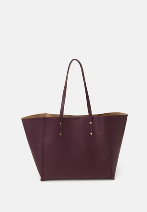 Bolso tote de cuero púrpura intenso con dos largas correas para el hombro y detalles de remaches de latón, lados inclinados, colocado sobre un fondo blanco liso.