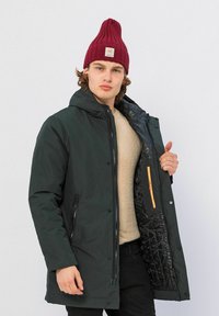 Donkergroen geïsoleerde jas met een capuchon, zwart doorgestikt interieur en oranje ritsdetails. Gedragen over een beige trui.