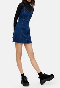 Denim dress with adjustable straps, structured bodice, and front button closure; paired with black platform sneakers and ribbed turtleneck.