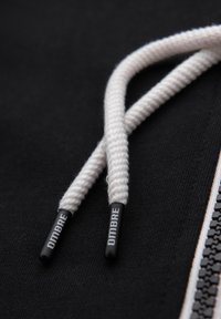 Cordon tressé blanc avec embouts en plastique noir portant l'inscription « OMBRE » sur un tissu noir près d'une fermeture éclair noire et blanche.