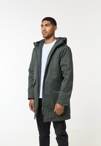 Zelená parka s kapucňou, ktorá má límec s fleece podšívkou, predné vrecká a hladký povrch. Nosená cez biele tričko a čierne nohavice.