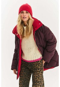 Bordowa kurtka puchowa z czerwonym kołnierzem, kremowy sweter z dzianiny z różowym obszyciem oraz spodnie w panterkę. Jasnoczerwony wełniany kapelusz.