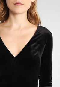 Svart sammetstopp med långa ärmar och djup V-ringning, med en mjuk textur och åtsittande design. Ingen synlig hårdvara eller mönster.