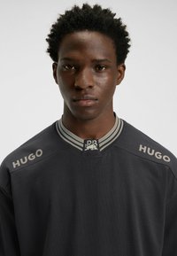 Sort sweatshirt med grå tekst "HUGO" på begge skuldre, en stribet krave og et logo-tag. Glat tekstur med en afslappet pasform.