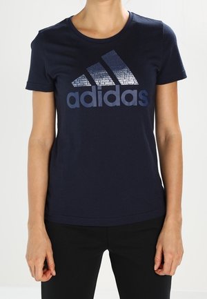 Frau trägt ein dunkelblaues Adidas T-Shirt mit großem Logo und Text auf der Brust, kombiniert mit schwarzen Hosen vor schlichtem Hintergrund.
