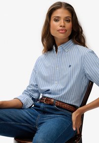 Kobieta siedząca na krześle, ubrana w koszulę na guziki w niebiesko-białe paski, z brązowym plecionym paskiem i niebieskimi dżinsami.