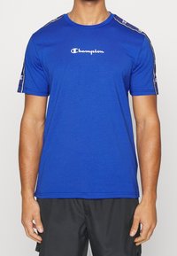 Modré sportovní tričko vyrobené z měkké tkaniny s krátkými rukávy, se bílým logem "Champion" na hrudi a vzorovanými akcenty na ramenech.