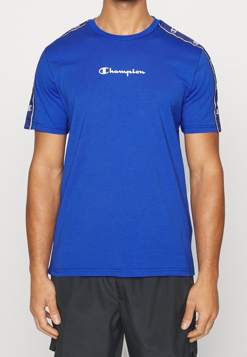 Modré sportovní tričko vyrobené z měkké tkaniny s krátkými rukávy, se bílým logem "Champion" na hrudi a vzorovanými akcenty na ramenech.