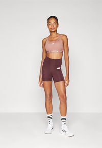 Atletisk antrekk som har en rosa sportsbh med hvit logo og brune shorts med hvite sidestreker. Teksturert stoff, tettsittende design.