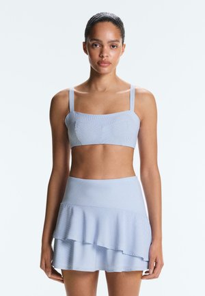 Žena nosi svijetloplavi prugasti crop top i odgovarajuću višeslojnu mini suknju, stoji na jednostavnoj bijeloj pozadini, okrenuta prema naprijed.