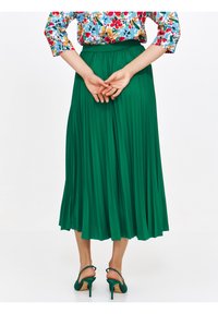 Zielona, plisowana spódnica midi z wysokim stanem i gładką fakturą. Noszona z pasującymi zielonymi szpilkami. Widoczna koszula w kwiatowy wzór.