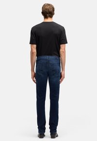 Męskie ciemnoniebieskie dżinsy o wąskim kroju, z pięcioma kieszeniami, kontrastującymi szwami i średnim stanem. Noszone z czarnym T-shirtem.