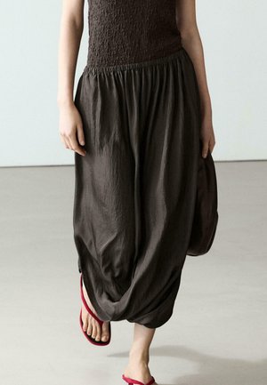 Donna che indossa una gonna larga marrone scuro con vita elastica e orlo arricciato, abbinata a sandali rossi con cinturini, cammina su un pavimento chiaro.