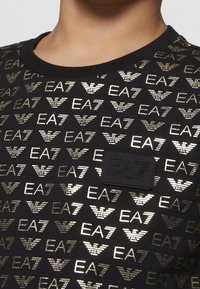 T-shirt in cotone nero con motivo logo EA7 dorato e toppa nera testurizzata con etichetta "EA7 Emporio Armani" sul petto.