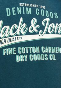 T-shirt in cotone verde acqua con testo bianco e verde chiaro. Il design include il nome del marchio, "Jack & Jones," e ulteriori dettagli grafici.