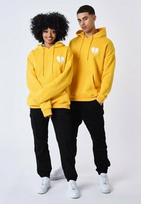 Sudaderas amarillas con un gráfico de corazón blanco, bolsillos tipo canguro en la parte delantera y cordones ajustables; combinadas con pantalones cargo negros y zapatillas blancas.