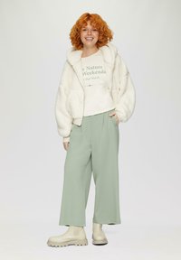 Lichte, zachte crème bomberjack over een lichtgroene sweatshirt met tekst, gecombineerd met groene, brede broekspijpen en beigekleurige enkellaarsjes.