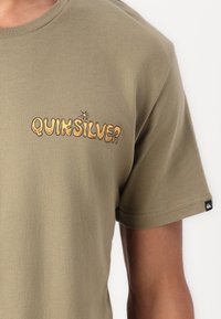 Alyvuogių spalvos medvilninis marškinėliai trumpomis rankovėmis, su geltonu ir juodu "QUIKSILVER" logotipu kairėje krūtinės pusėje. Pridedama maža prekės etiketė.