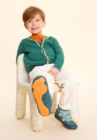 Cardigan verde a maglia con bordi arcobaleno, maglietta arancione, pantaloni crema e scarpe turchesi con due fasce in velcro e suola arancione, seduto su una sedia maculata.