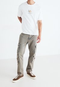 T-shirt bianca in cotone con grafica floreale, pantaloni cargo grigi con molte tasche laterali e sneakers in suede marrone con suole bianche.