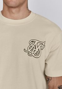 Helle beige Baumwoll-T-Shirt mit rundem Halsausschnitt und kurzen Ärmeln; verfügt über ein schwarzes stilisiertes Logo im linken Brustbereich.