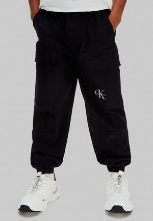 Person trägt schwarze Cargo-Hose mit Calvin Klein-Logo und weiße Sneaker mit sichtbaren Schnürsenkeln, steht mit den Händen in den Taschen.