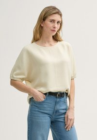 Cremfarbener Kurzarmoberteil mit strukturierter Oberfläche, Rundhalsausschnitt und lockerer Passform, kombiniert mit hellblauen hochgeschnittenen Jeans und einem schwarzen Gürtel.