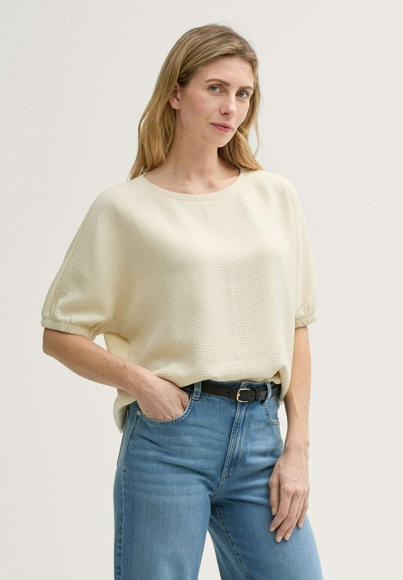 Cremfarbener Kurzarmoberteil mit strukturierter Oberfläche, Rundhalsausschnitt und lockerer Passform, kombiniert mit hellblauen hochgeschnittenen Jeans und einem schwarzen Gürtel.
