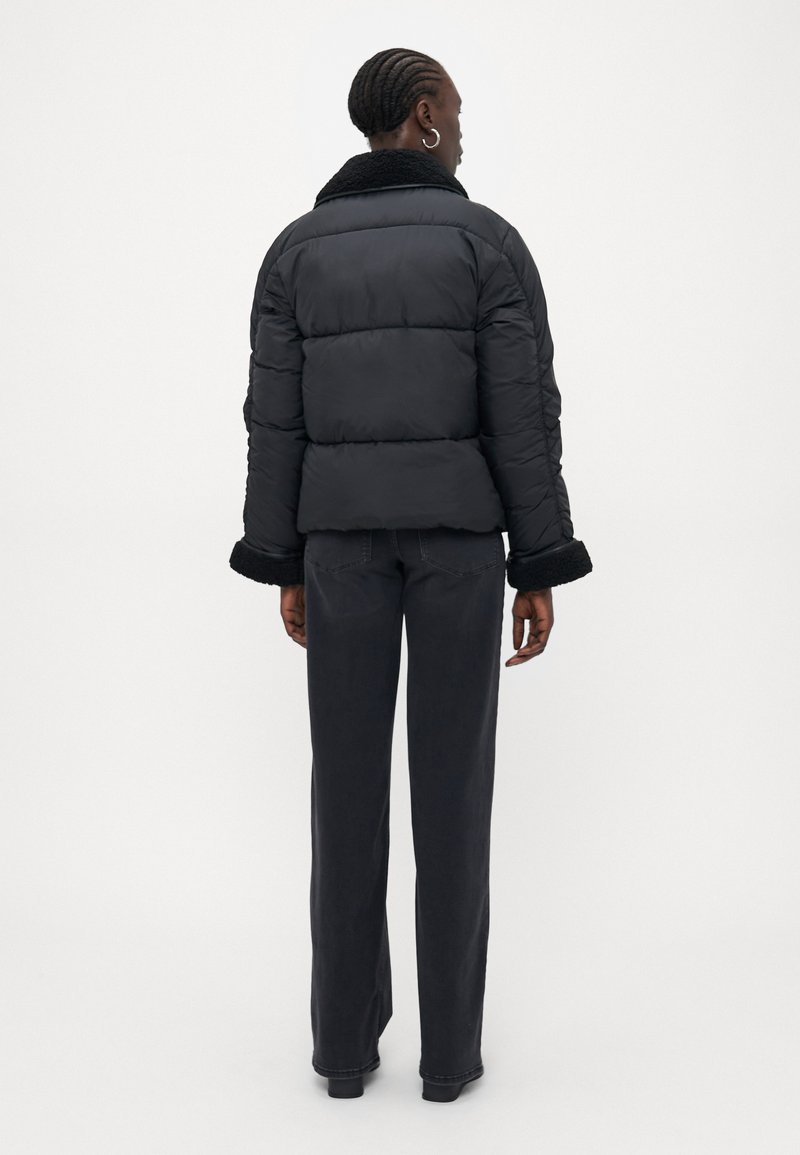 Giubbotto imbottito nero con colletto e polsini in peluche, caratterizzato da una texture a righe orizzontali. Indossato con jeans a gamba larga scuri.