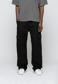 Person in schwarzen Cargo-Hosen mit großen Seitentaschen, einem verblassten grauen T-Shirt und weißen Sneakers vor einem schlichten grauen Hintergrund.