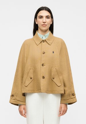 Γυναίκα που φοράει μπεζ κάπα-μπουφάν με κουμπιά και μπροστινές τσέπες πάνω από ένα ανοιχτοπράσινο πουκάμισο με γιακά και λευκό παντελόνι, όρθια μπροστά σε απλό φόντο.