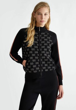 Cardigan nero con zip, caratterizzato da un motivo ripetitivo, polsini a costine e strisce laterali in rosso e beige. Texture liscia, design aderente.