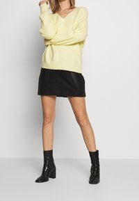 Gelber Strickpullover mit V-Ausschnitt, kombiniert mit einem schwarzen Minirock aus Kunstleder und glänzenden schwarzen Knöchelstiefeln mit Blockabsatz.