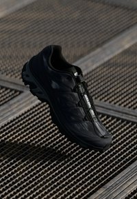 Black Salomon trail running shoe resting on a metal grate surface, showing side and top with textured sole and quick lace system.