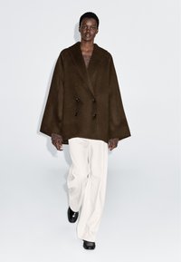 Manteau marron à double boutonnage avec une coupe ample, fabriqué en laine, doté de grands boutons noirs et de larges manches, associé à un pantalon blanc.