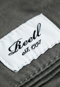 Weißes Stoffetikett mit schwarzer kursiver Schrift "Reell" und "est. 1997", auf einem grauen strukturierten Material angenäht, das detaillierte Stickarbeiten zeigt.