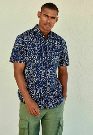 Kurze Ärmel Hemd in Marineblau mit einem weißen Blumenmuster; kombiniert mit hellgrünen Cargo-Hosen mit seitlichen Taschen und lockerer Passform.