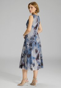 Un vestido sin mangas, fluido, en tono gris claro con un patrón floral abstracto en azul y marrón. La modelo lleva sandalias plateadas de tiras.