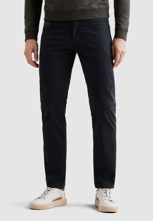 Mand iført mørkeblå slim-fit bukser, hvide sneakers med beige såler og en mørkeolivengrøn langærmet top, stående mod en ensfarvet baggrund.