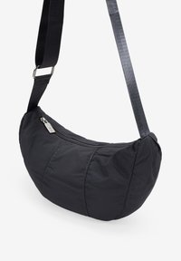 Bolso cruzado negro de nailon con un diseño redondeado, cierre de cremallera y correa ajustable. Costuras visibles y un sutil detalle de brillo.